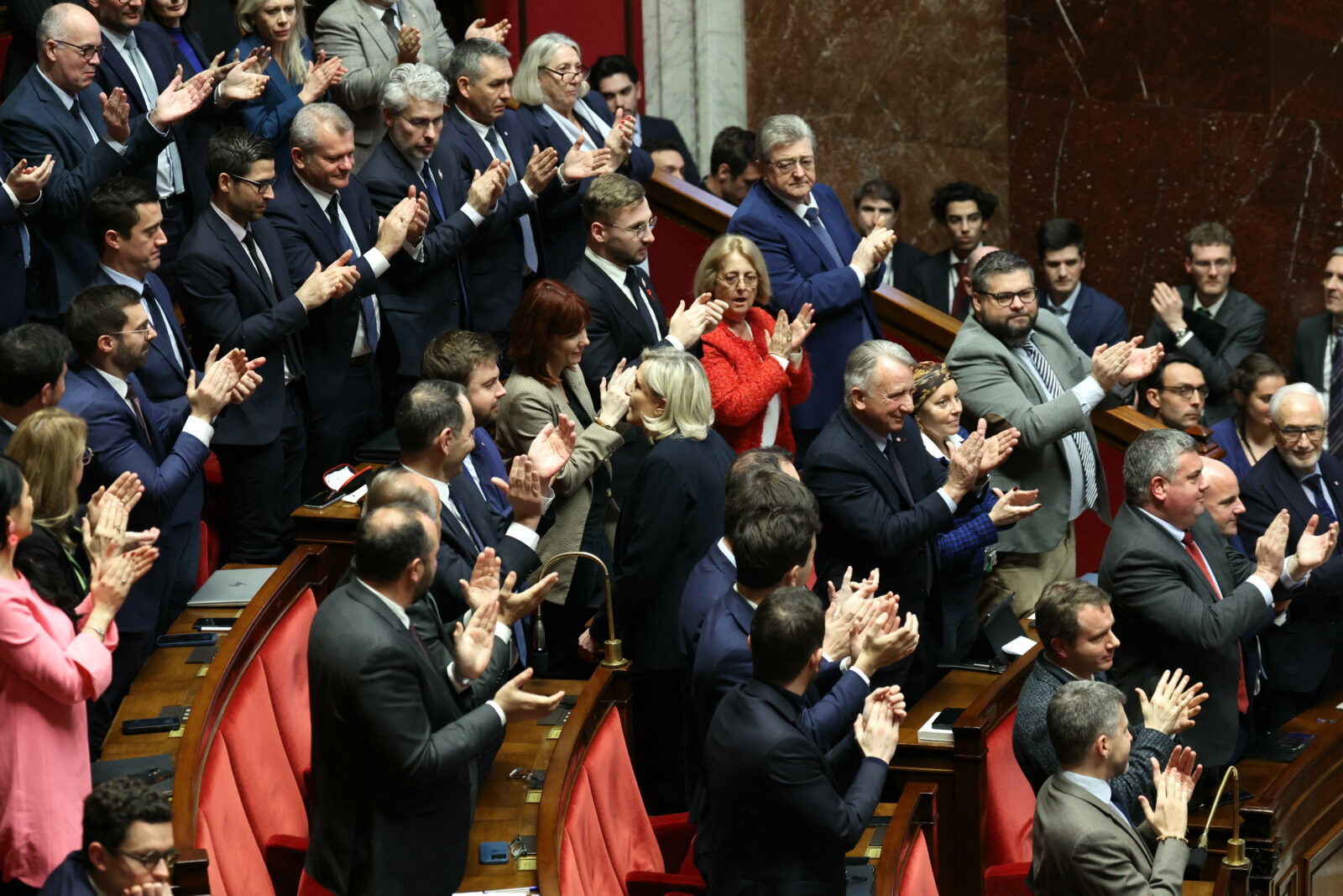 President of Rassemblement National parliamentary group Marine Le Pen (C), applauded by French far-right Rassemblement National (National Rally) RN's parliamentary group, reaches her seat after she delivered her speech during the debate prior to the no-confidence votes on Prime Minister Michel Barnier's administration at the National Assembly in Paris on December 4, 2024. - The French National Assembly debates two motions brought by the French left-wing Nouveau Front Populaire (New Popular Front) NFP coalition and the French far-right Rassemblement National (National Rally) RN party in a standoff over 2025's austerity budget, which saw French Prime Minister force through a social security financing bill without a vote (article 49.3) on December 2, 2024. (Photo by Alain JOCARD / AFP)<img src=