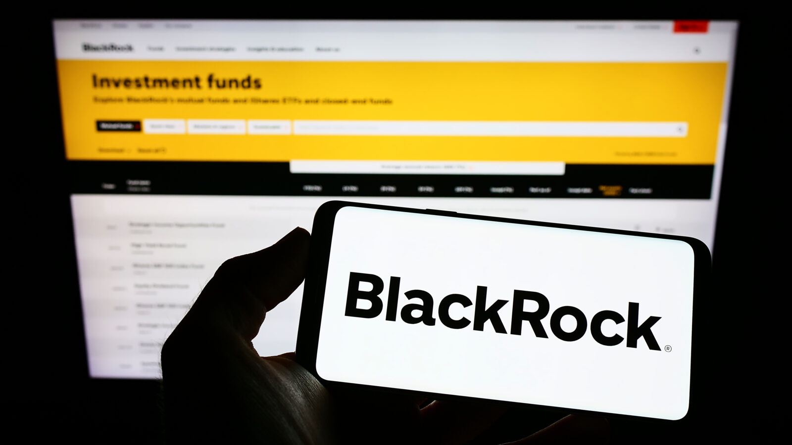 The file photo shows a person holding a smartphone displaying the BlackRock logo in front of a computer monitor showing the BlackRock Investment Funds webpage. (Adobe Stock Photo)