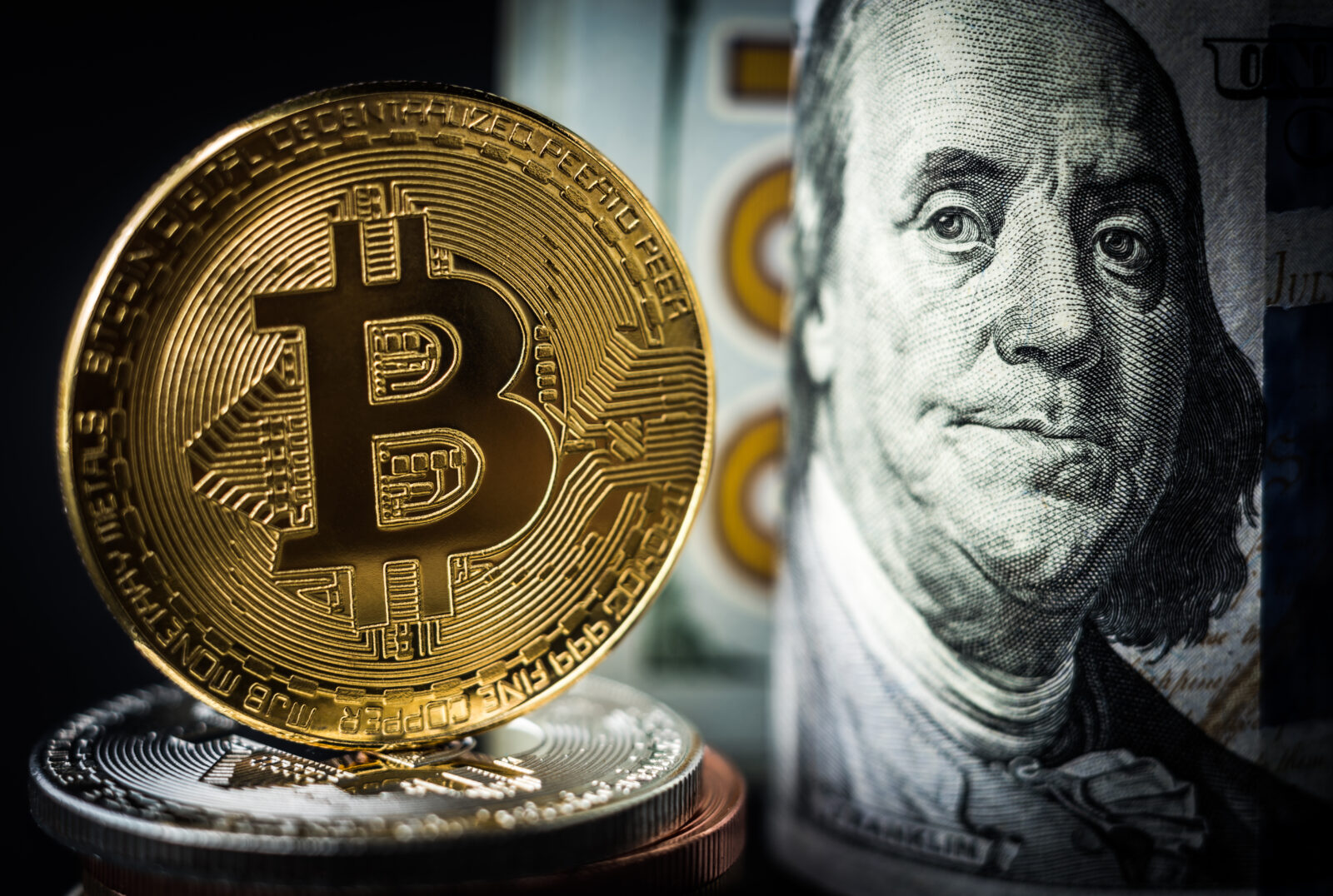 A close-up image of a physical Bitcoin coin stacked on other cryptocurrency coins, with a rolled-up U.S. dollar bill in the background. (Adobe Stock Photo)