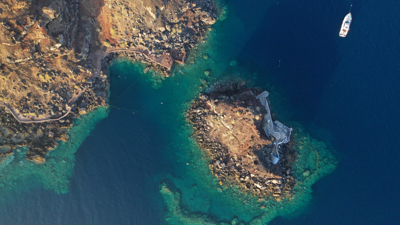 Aerial drone photo of picturesque volcanic islet with black rocks and the Chapel of Agios Nikolaos in the bay of Amoudi beneath the iconic village of Oia, Santorini, Greece. (Adobe Stock Photo)