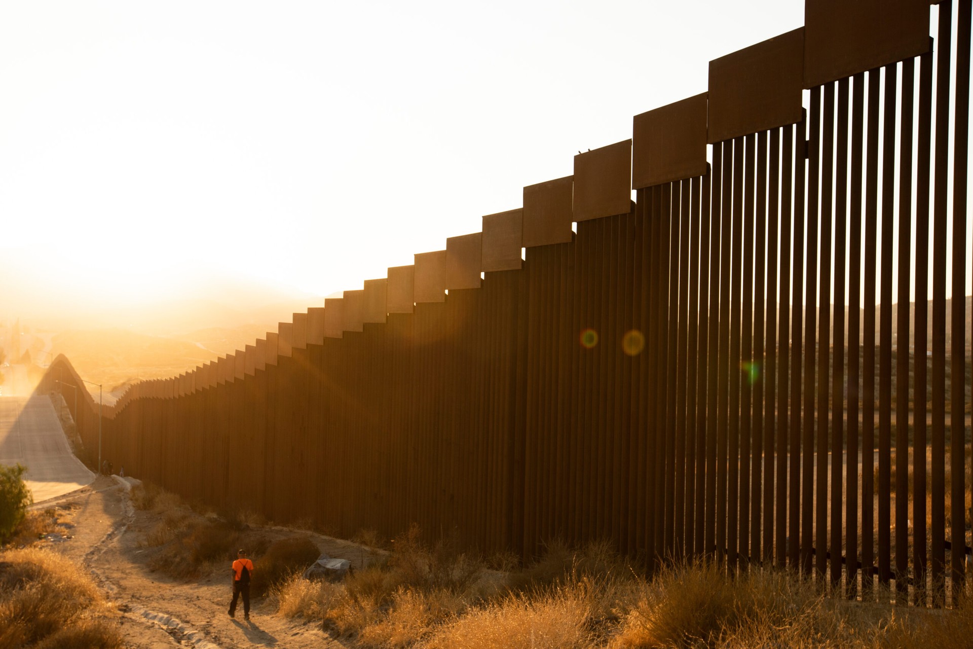 A person walks along a dusty path beside the towering steel barrier of the U.S.-Mexico border wall in Tecate, Baja California, on Sep. 14, 2021. (Adobe Stock Photo)