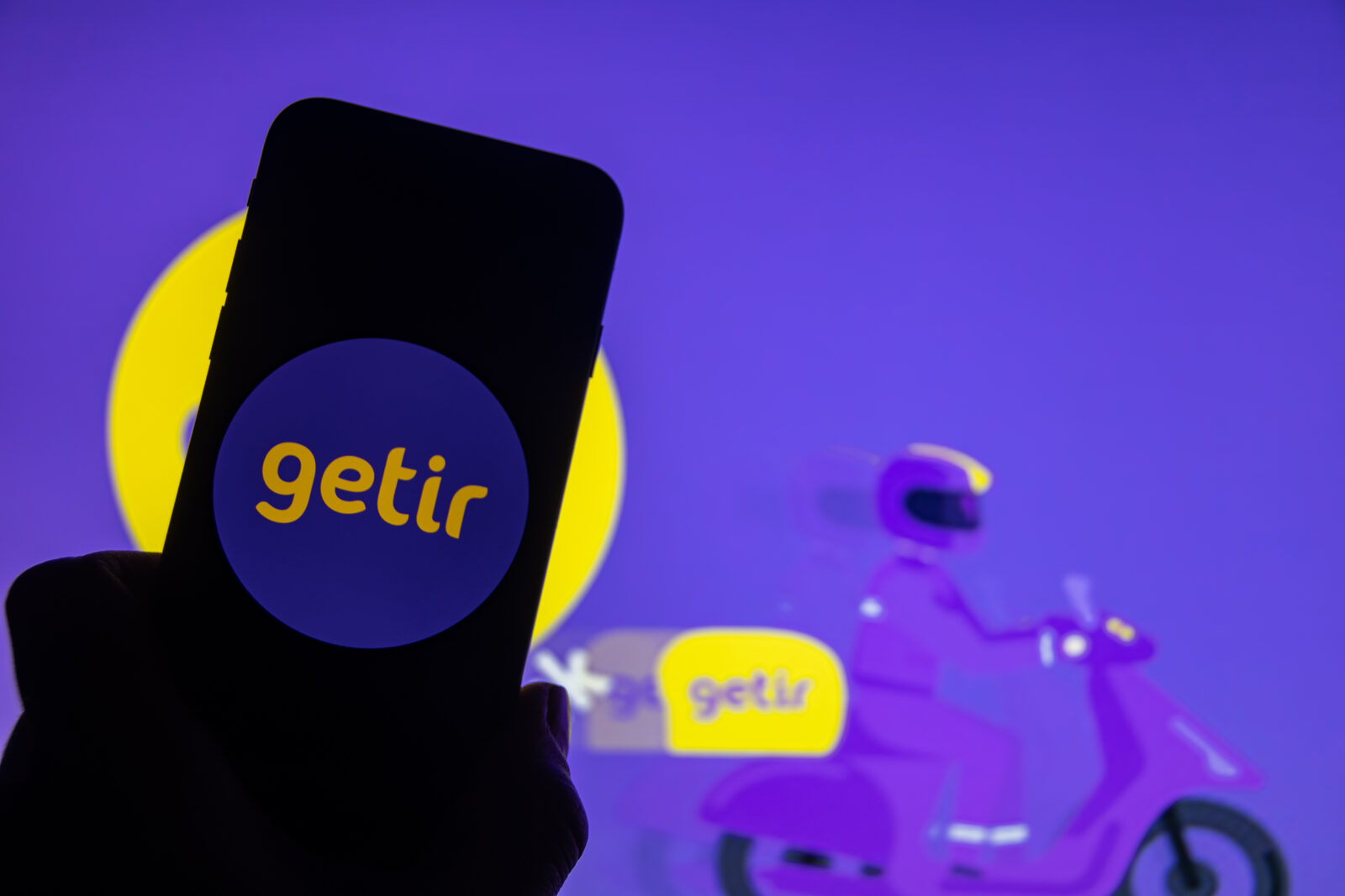 The file photo shows a smartphone displaying the Getir logo held up against a blurred background featuring a Getir delivery rider on a scooter. (Adobe Stock Photo)