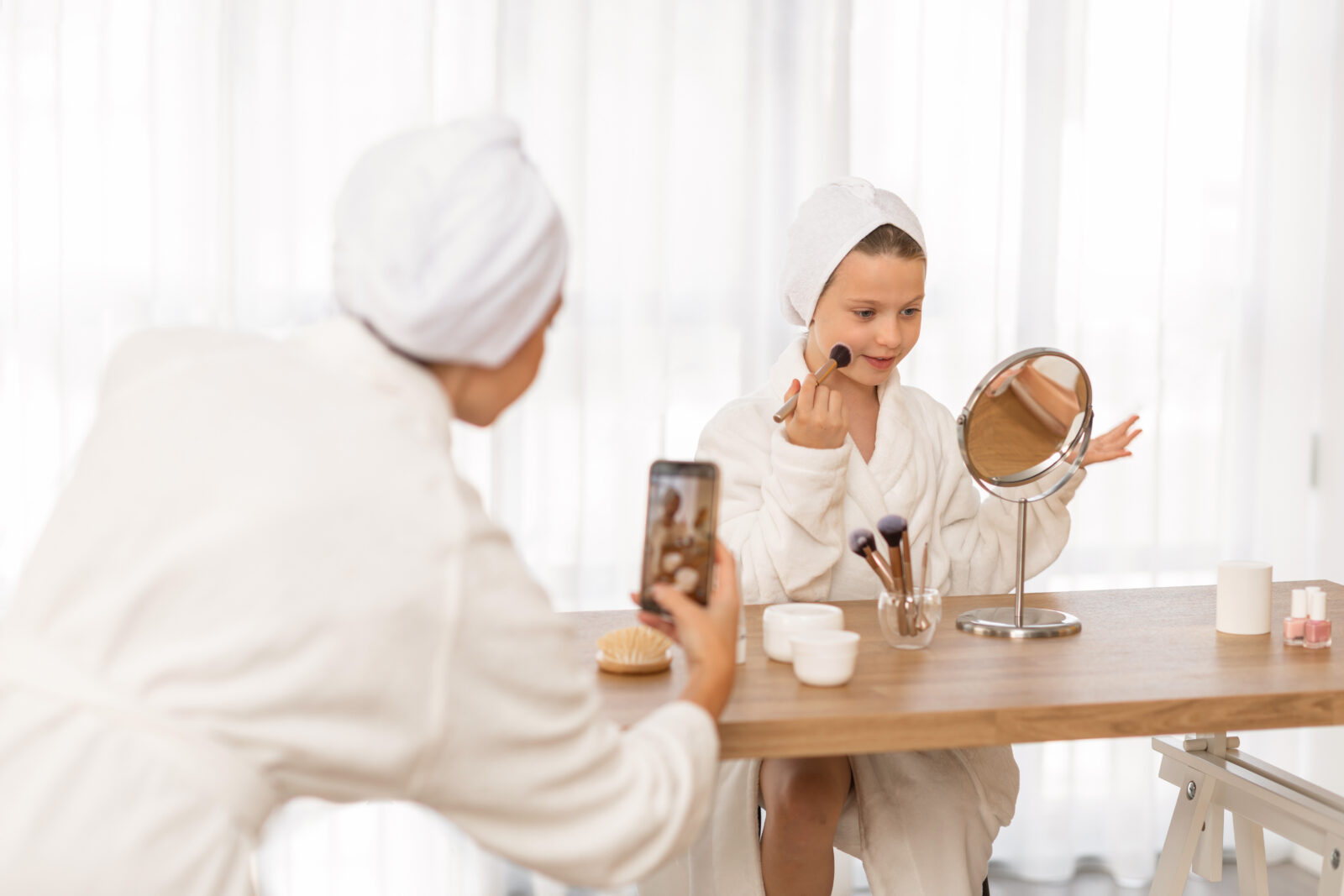 A little girl and her mom, both wearing bathrobes, record a makeup tutorial at home. The adorable child applies makeup with a brush while her mother captures the moment for social media using a smartphone. (Adobe Stock Photo)