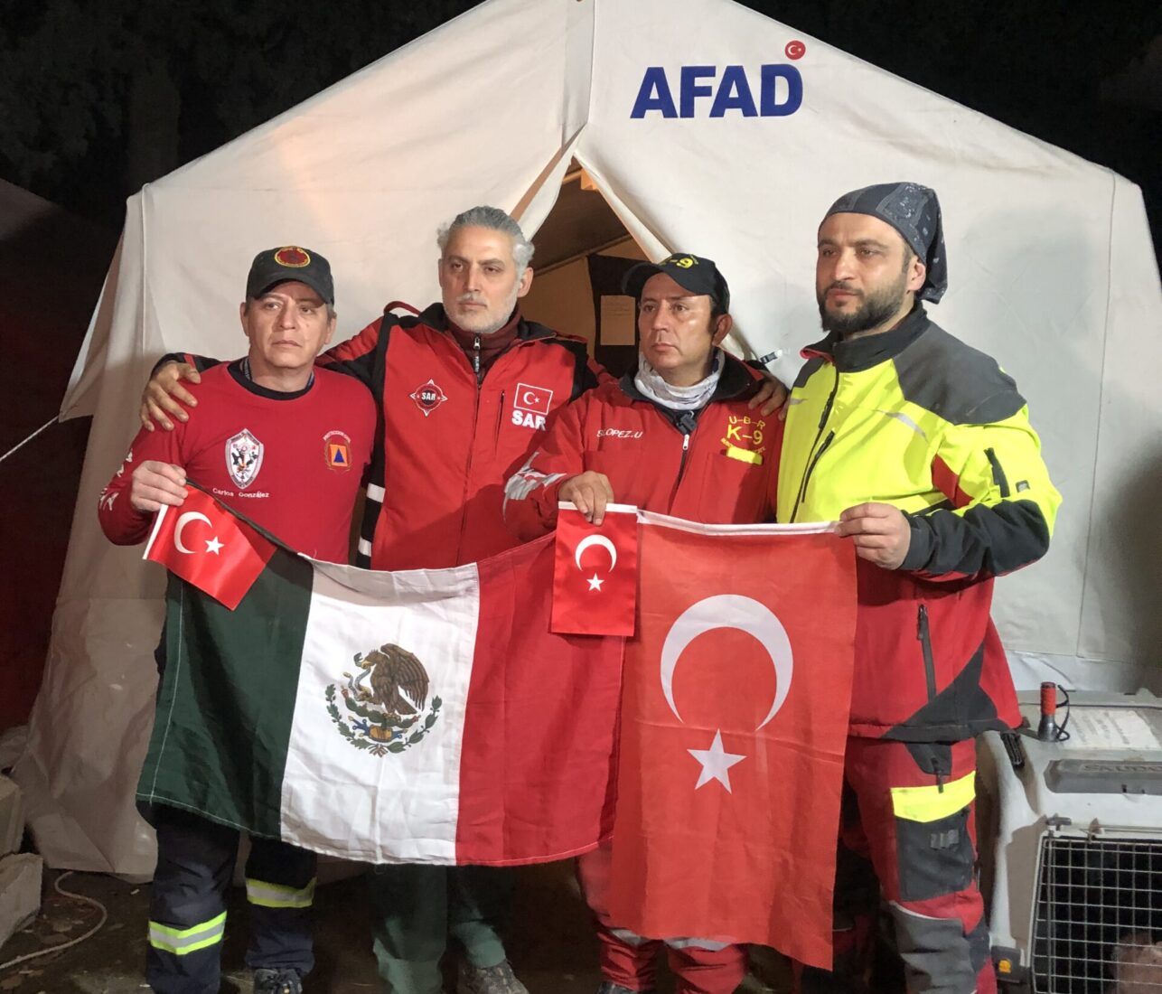 Mexican volunteers and Turkish SAR Organization president Erdem Veysel Akin hold their national flags in front of an AFAD tent during rescue efforts for the 2023 earthquakes, Antakya, Türkiye, February 2023. (Courtesy of SAR)