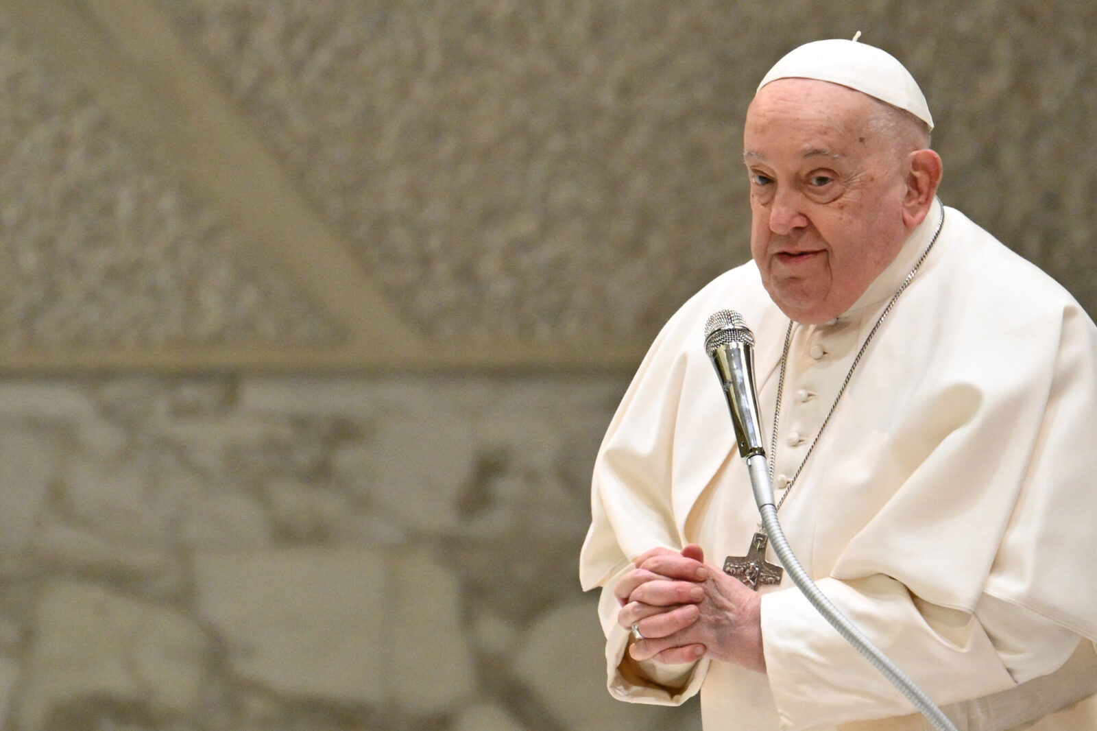 Pope Francis, dressed in white, speaks into a microphone against a textured stone background. (Photo via X)