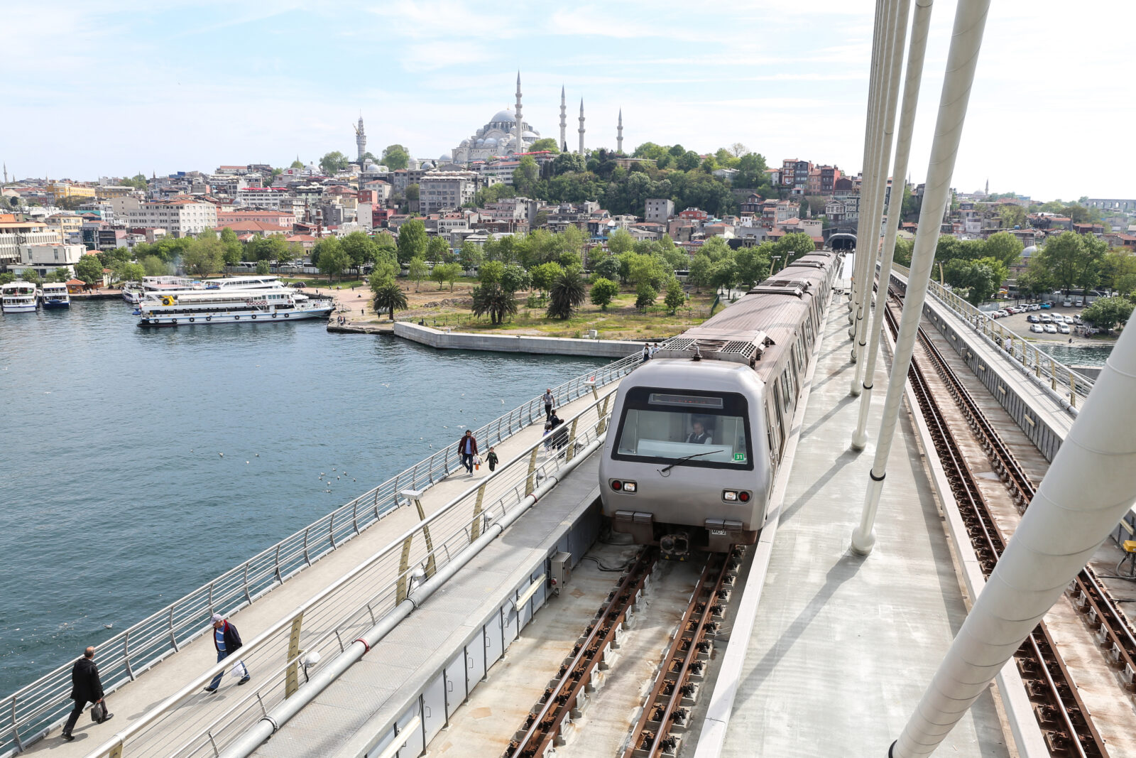 A metro train crosses the Golden Horn Metro Bridge in Istanbul with the Suleymaniye Mosque in the background, as locals and tourists walk along the pedestrian path on a clear spring day, Türkiye. (Adobe Stock Photo)