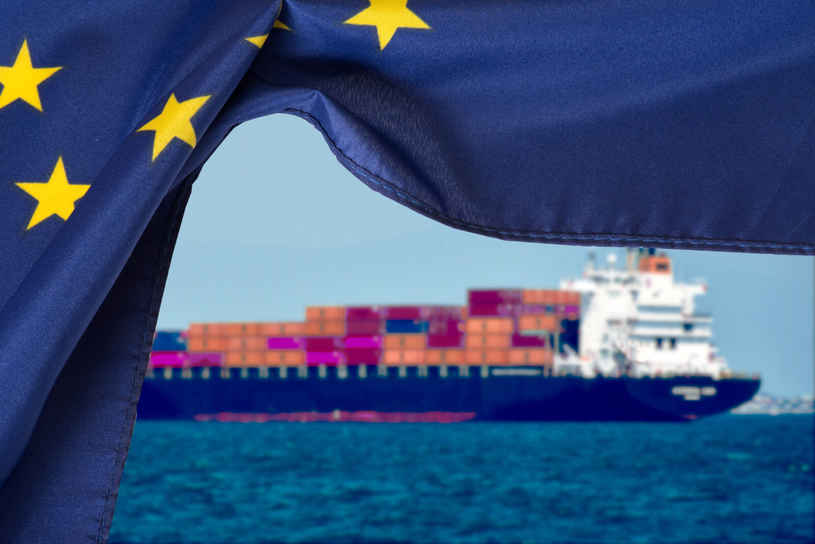 The European Union flag waves in the foreground as a container ship navigates waters, accessed on March 12, 2025. (Adobe Stock Photo)