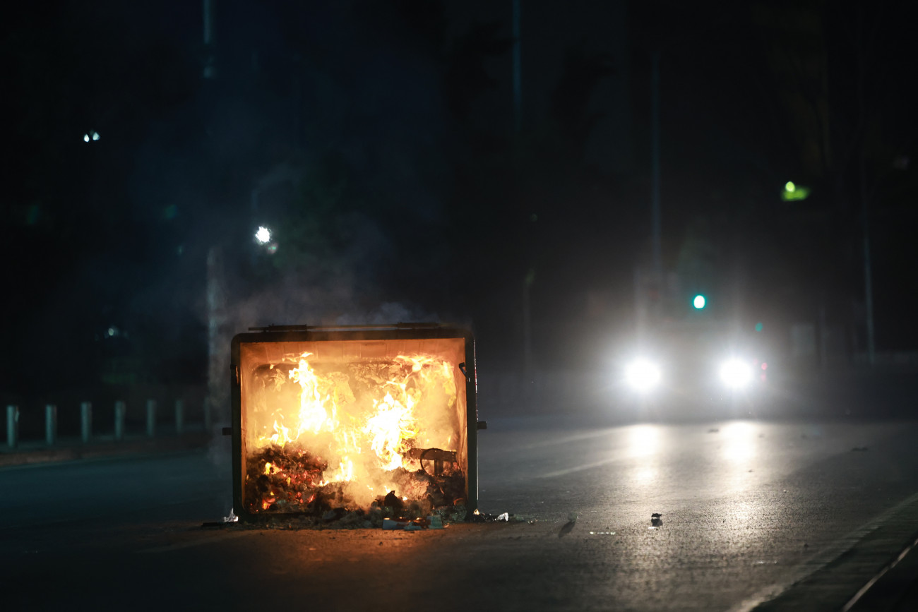 A burning trash bin blocks a road at night, with car headlights illuminating the scene, during protests in Athens over the Tempi train accident.