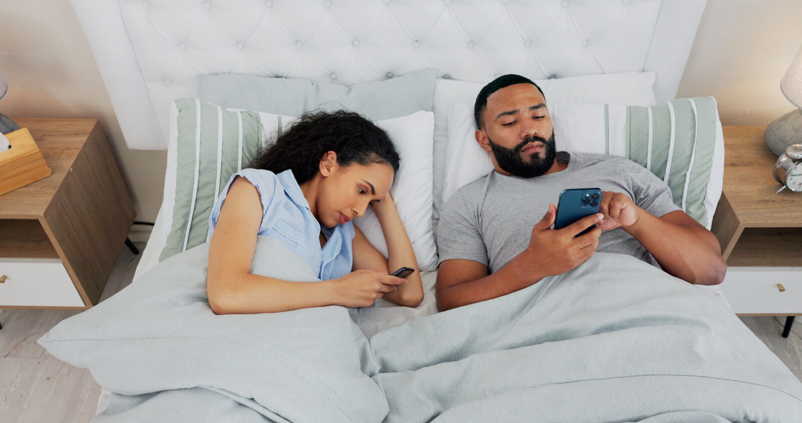 A couple lying in bed side by side, both absorbed in their smartphones rather than engaging with each other, highlighting digital distraction in intimate relationships, accessed on April 26, 2025. (Adobe Stock Photo)
