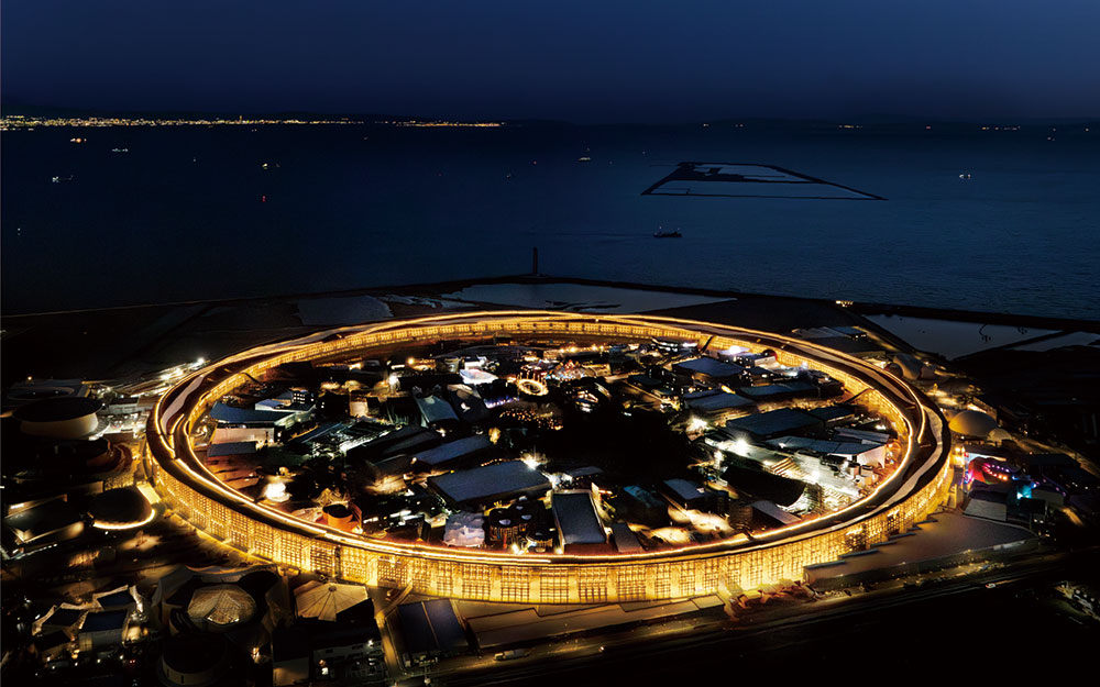 Aerial night view of the Grand Ring at Expo 2025 Osaka illuminated against Osaka Bay, showcasing the circular exhibition hub's golden glow and intricate architectural design, accessed on April 13, 2025. (Photo via EXPO 2025)