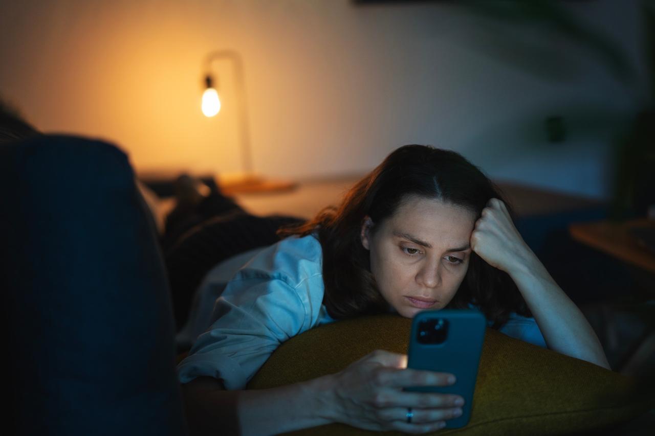 A woman in dim lighting looking stressed while checking her phone late at night, exhibiting signs of digital anxiety or doom-scrolling behavior, accessed on April 26, 2025. (Adobe Stock Photo)