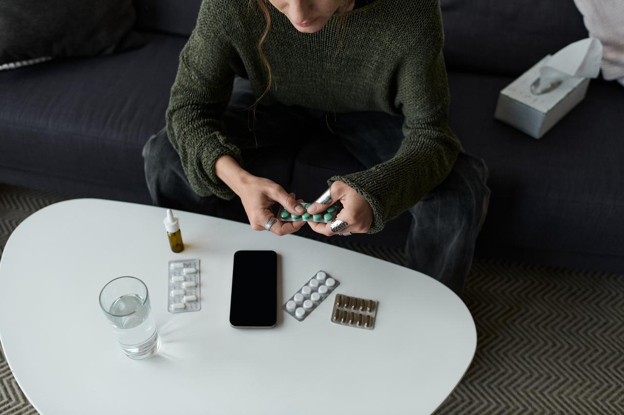 A person holding a pack of medication while sitting on a sofa, with pills, a glass of water, and a smartphone placed on a white table. (Adobe Stock Photo)