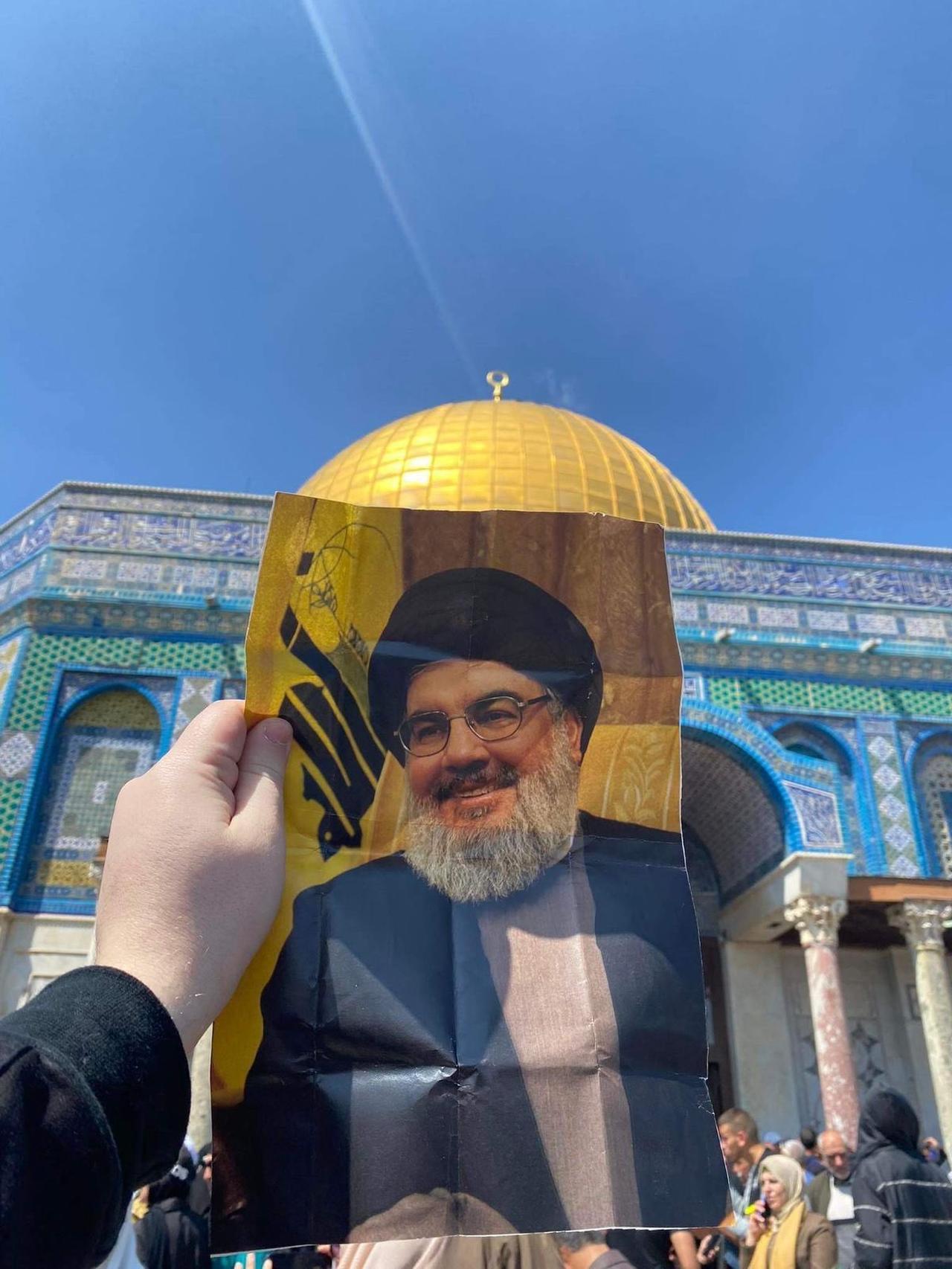 The image shows a person holding up a picture of Hassan Nasrallah, the former Secretary-General of Hezbollah, in front of the Dome of the Rock in Jerusalem.