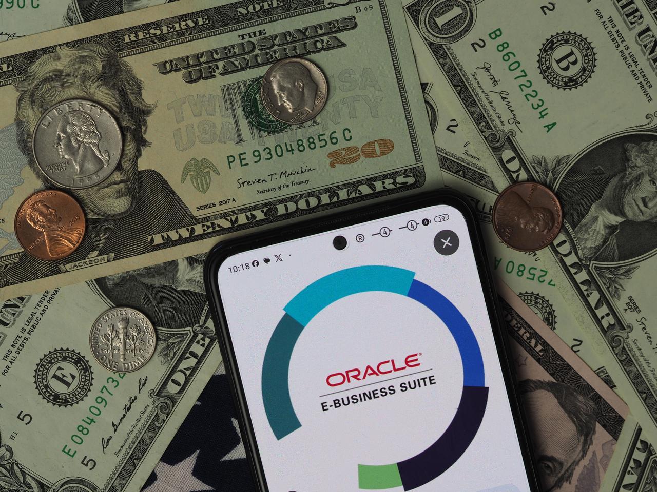 The Oracle E-Business Suite logo is seen displayed on a smartphone screen placed over U.S. dollar banknotes. (Adobe Stock Photo)