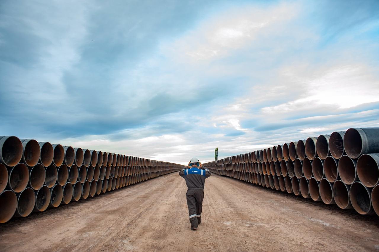 The image displays the construction site of the Vaca Muerta Sur Oil Pipeline (VMOS) in Argentina, featuring large diameter pipes and a worker. (Photo via YPF)