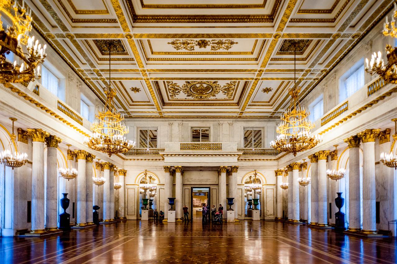 Visitors walk through a grand state hall of the Hermitage Museum, where marble columns, gilded chandeliers and a richly decorated ceiling recall the buildings imperial past, Feb. 24, 2015. (Adobe Stock Photo)