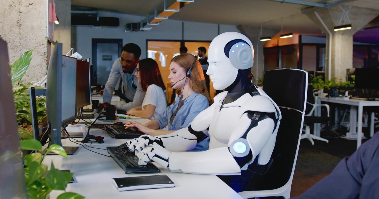 A robot using a computer alongside humans, symbolizing artificial intelligence. (Adobe Stock Photo)
