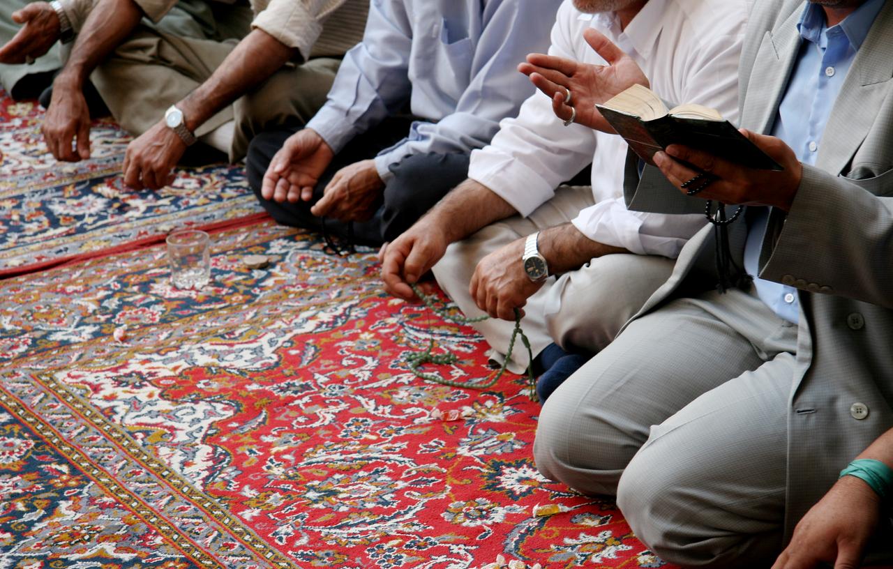 Men sit in prayer on a patterned mosque carpet during a communal gathering, with prayer beads and an open book visible. (Adobe Stock Photo)