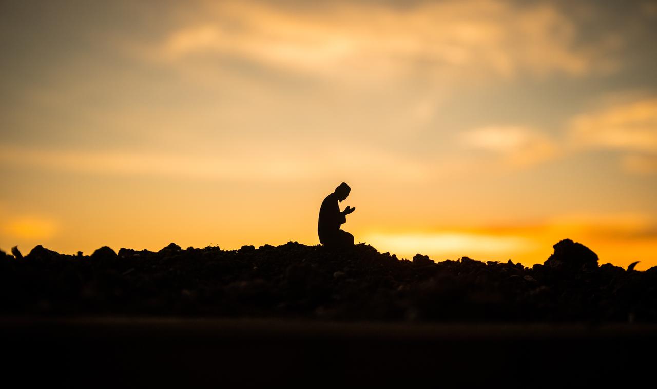 A man prays at sunset in a quiet outdoor setting, silhouetted against the sky. (Adobe Stock Photo)