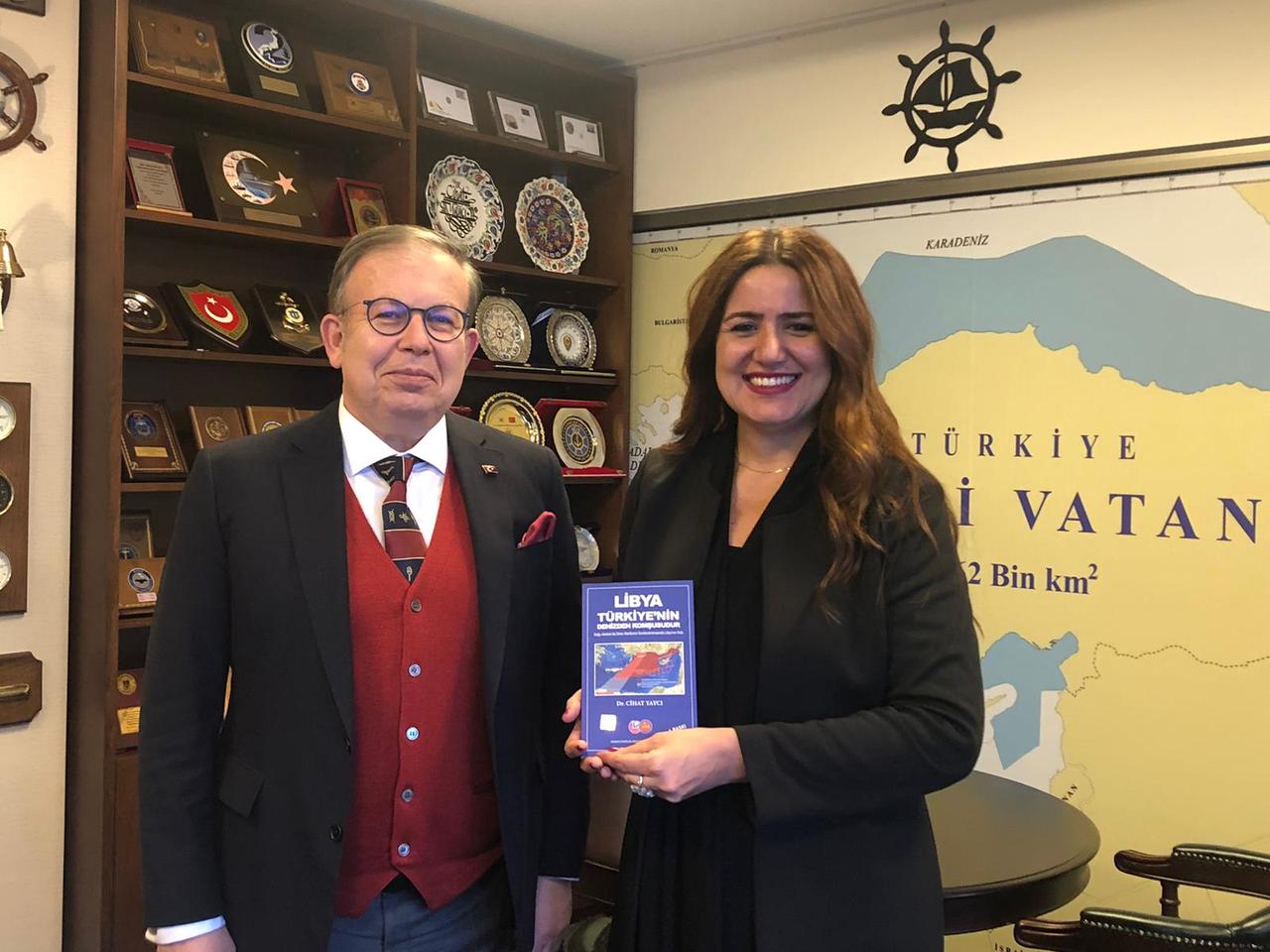 The image shows Cihat Yayci (L) and Elif Ulug. They are holding a book titled "Libya is Türkiye's Maritime Neighbor." The background features a map of 'Blue Homeland' doctrine.