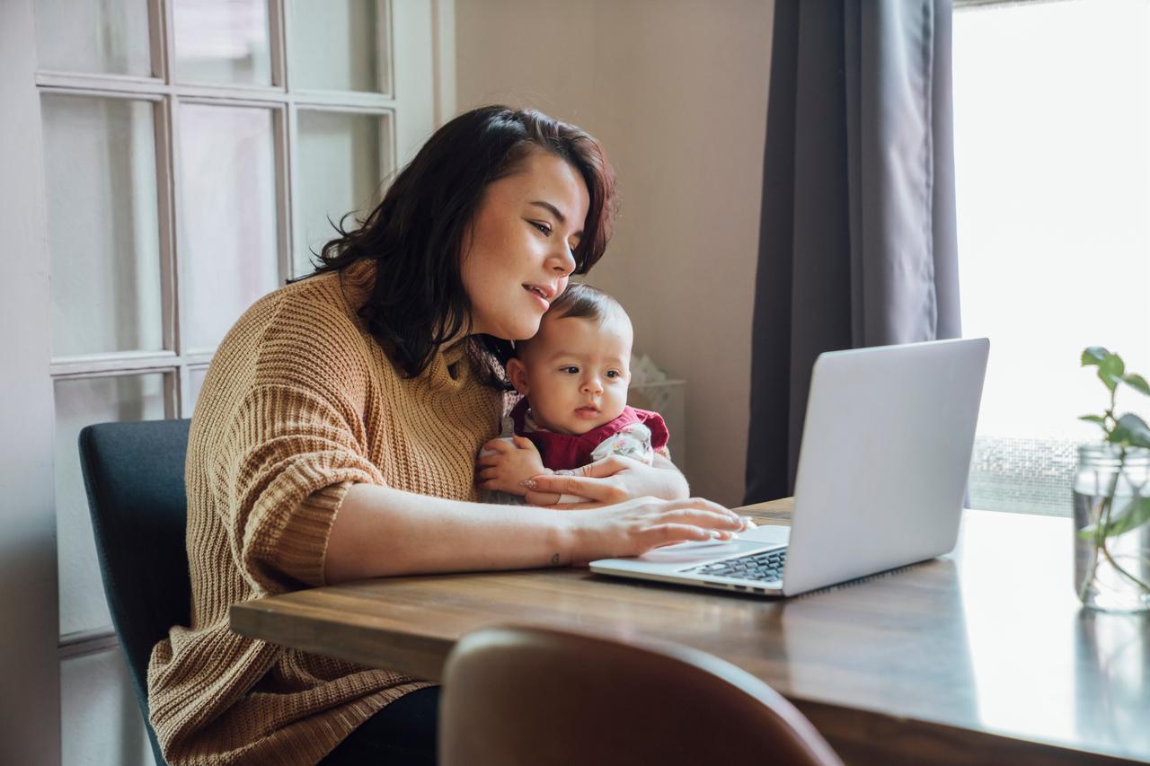 This image shows a person with a baby using a laptop computer at a table. (Adobe Stock Photo)