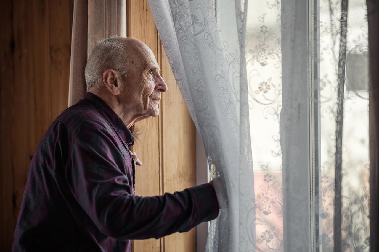 Single elderly man looking through window glass standing at home. (Adobe Stock Photo)