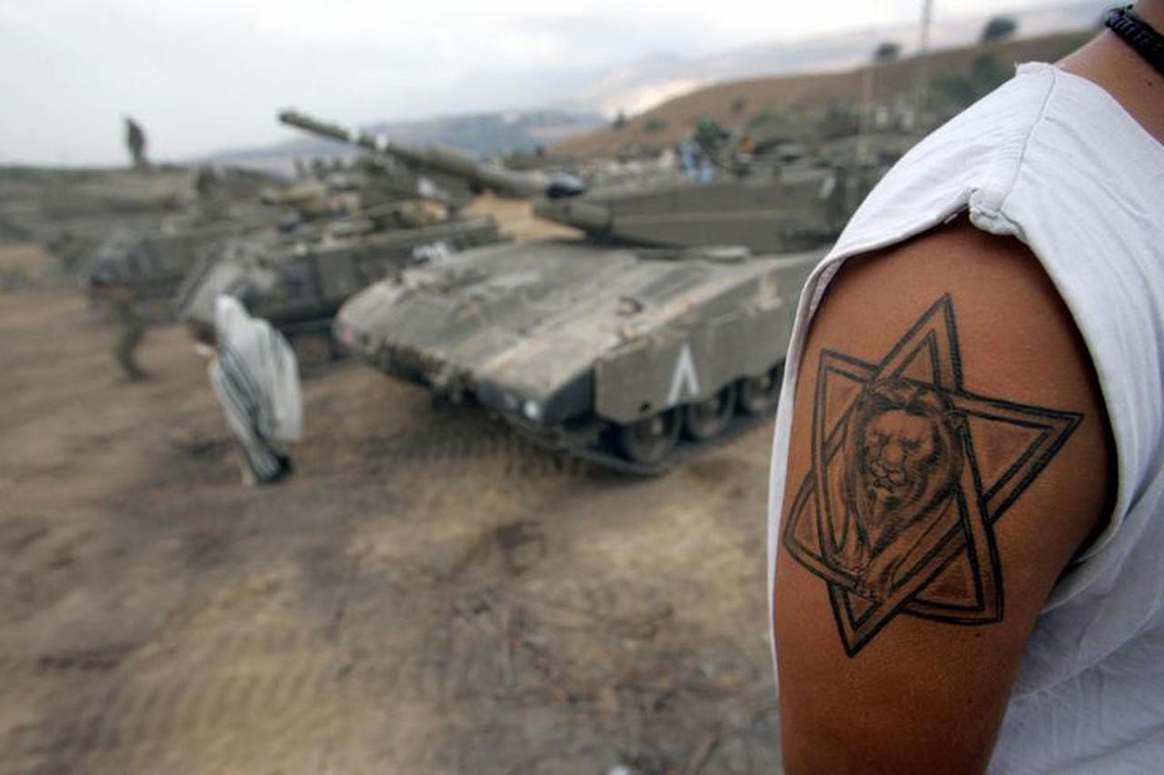 An Israeli army reservist, who sports a tattoo of the Lion of Judah surrounded by a Star of David, works on his tank as another performs his morning prayers at a forward base where the Israeli army have deployed dozens of tanks and armored vehicles August 8, 2006 on Israel's border with Lebanon. (AFP Photo)