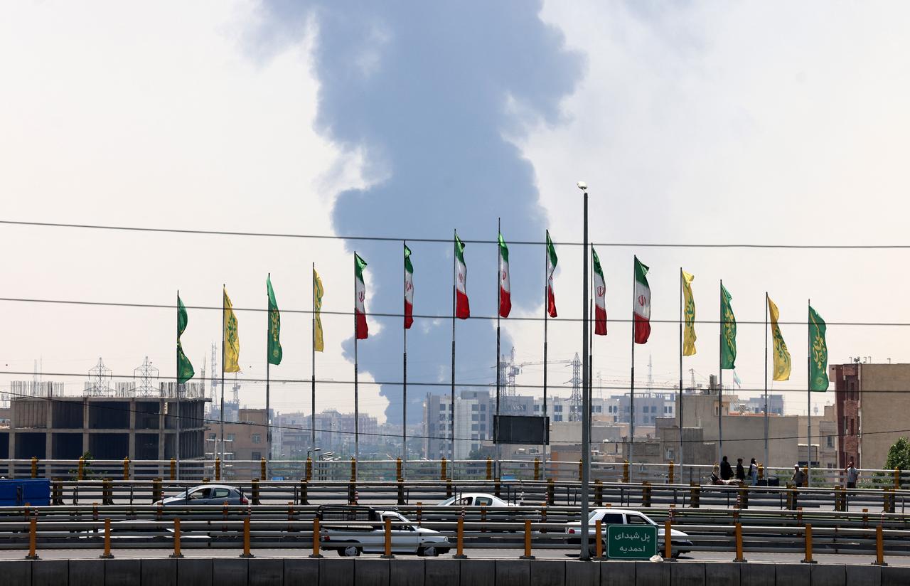 Flags flutter along a bridge as a plume of heavy smoke and fire rise from an oil refinery in southern Tehran, after it was hit in an overnight Israeli strike, on June 15, 2025. (AFP Photo)