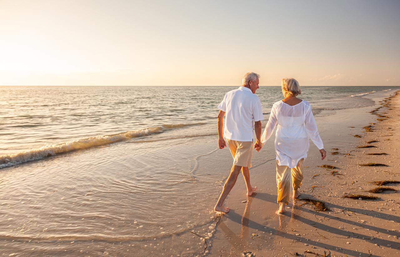 Elderly retired couple strolling hand in hand along the beach at sunset. (Adobe Stock Photo)