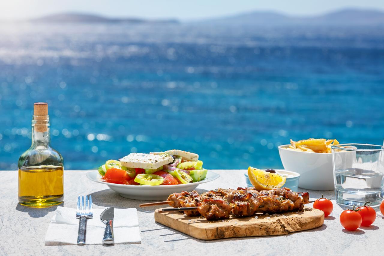Greek cuisine with a farmers salad and souvlaki skewers-a dish also deeply rooted in Turkish culinary tradition-set against the sparkling blue Aegean Sea in summer. (Adobe Stock Photo)
