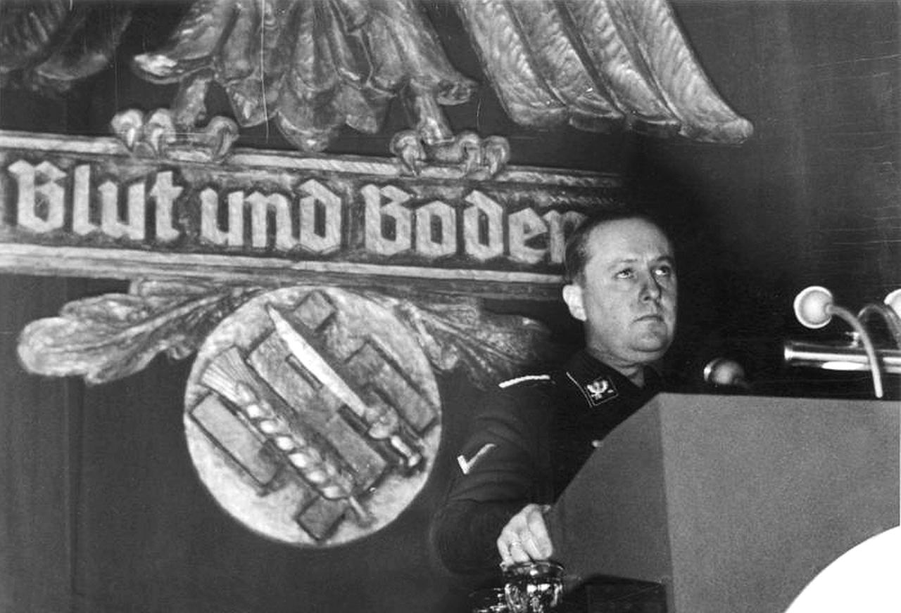 Richard Walther Darré addresses a farming community meeting in Goslar on Dec. 13, 1937, standing before a Reichsadler and swastika emblem crossed with a sword and wheat sheaf, symbolising the Nazi slogan "Blood and Soil." (Photo via German Federal Archive)