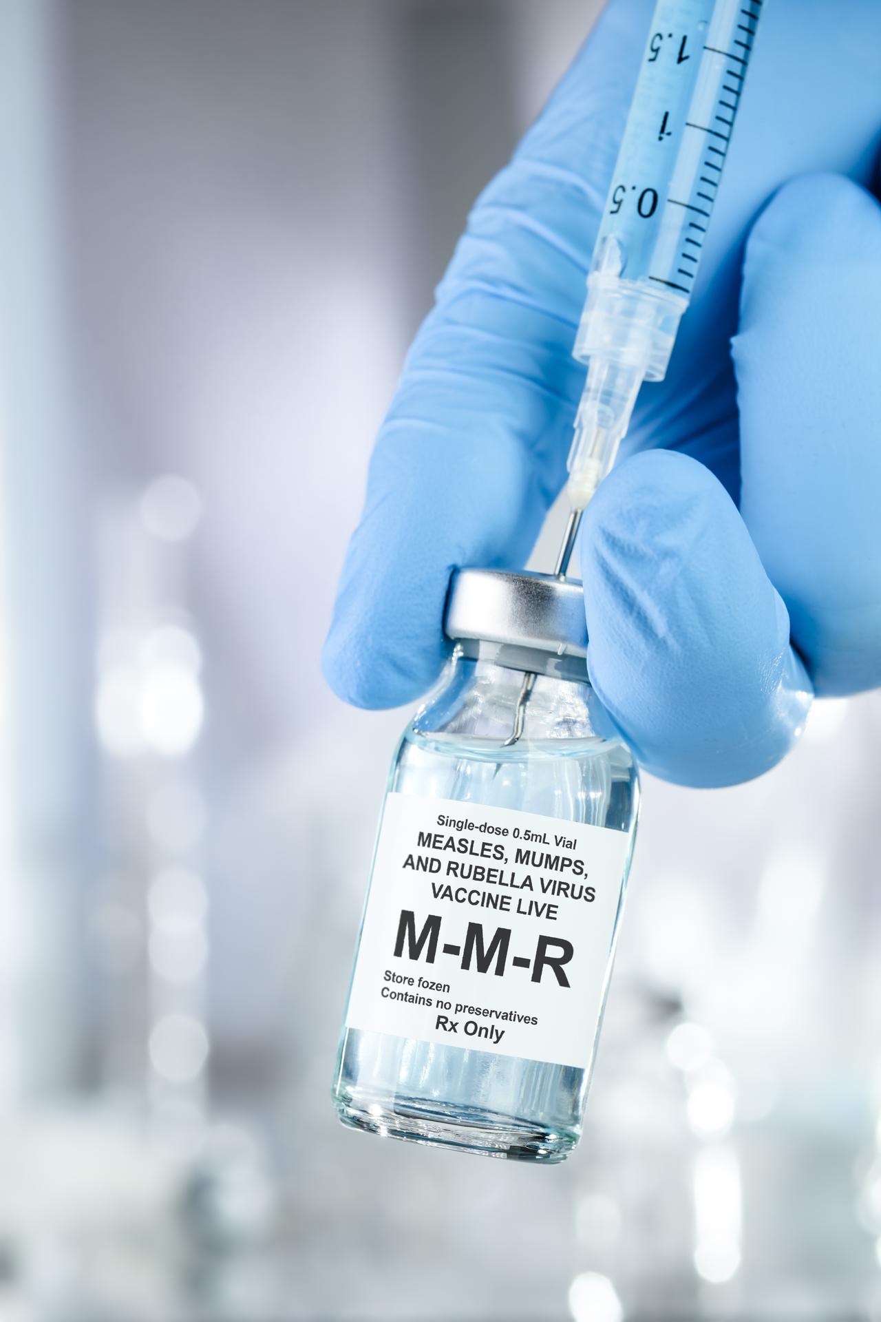 A person wearing blue medical gloves is holding a vial of the MMR vaccine (measles, mumps, and rubella) along with a syringe and needle. (Adobe Stock Photo)
