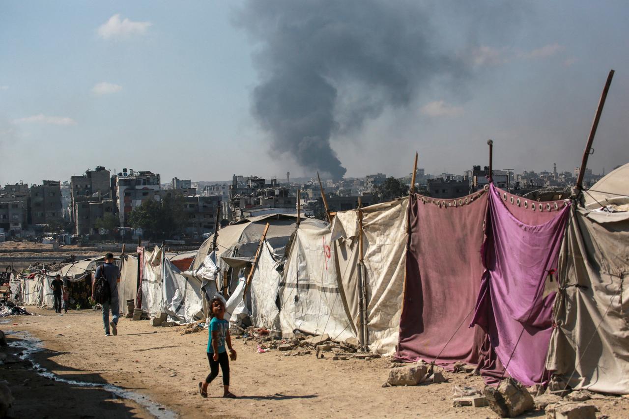 People walk past makesfift shelters as smoke billows east of Gaza City, in the central Gaza Strip during Israeli bombardment on July 6, 2025. (AFP Photo)