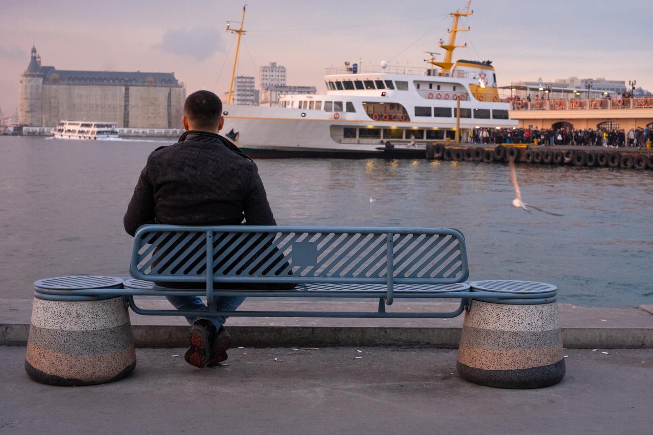 A man sits alone on a bench by the waterfront, watching the ferries come and go as evening falls over Istanbul in Türkiye. (Adobe Stock Photo)