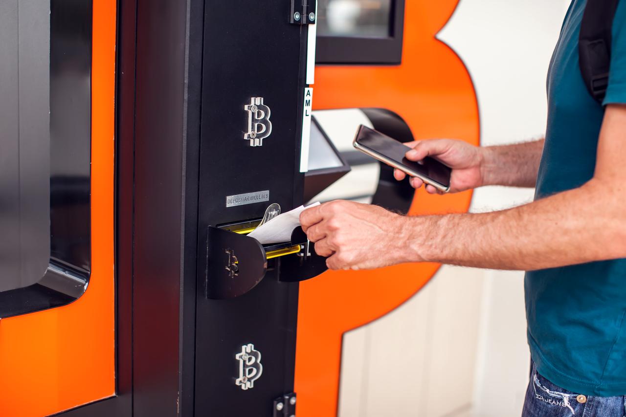 A man uses a Bitcoin ATM to deposit cash while holding his smartphone. (Adobe Stock Photo)