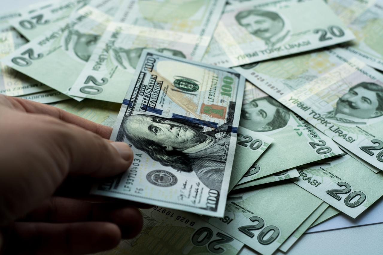 A person holds a U.S. dollar banknote over a pile of Turkish lira notes. (Adobe Stock Photo)