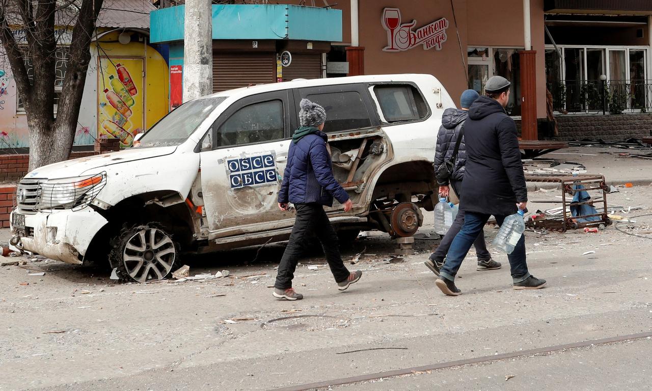 People walk past an OSCE car damaged in the course of Ukraine-Russia conflict in the southern port city of Mariupol, Ukraine, April 1, 2022. (Photo via TRT News)