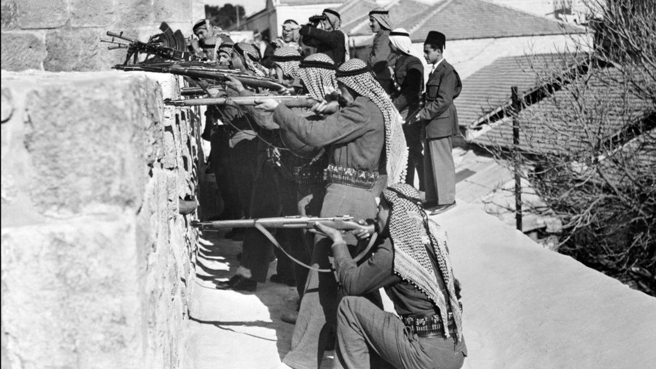 Armed men wearing traditional keffiyehs aim rifles over a stone wall, seemingly defending or attacking a position, while a few men in suits observe from behind, in what appears to be a historical black-and-white photo in an urban setting, accessed on Sept. 20, 2025. (AFP Photo)