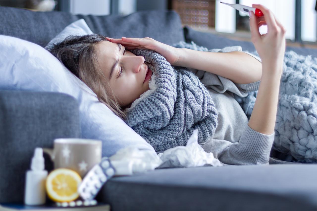 A young woman with a headache and fever checks her temperature using a thermometer. (Adobe Stock Photo)