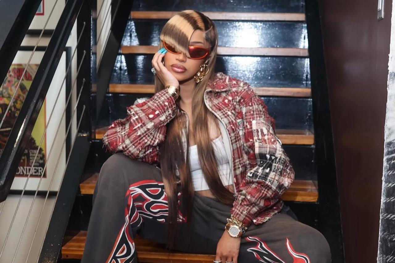 Cardi B poses on a staircase, wearing flame-print pants, layered gold jewelry, and holding an orange Birkin bag, Aug. 2, 2025. (Photo via Instagram/@iamcardib)