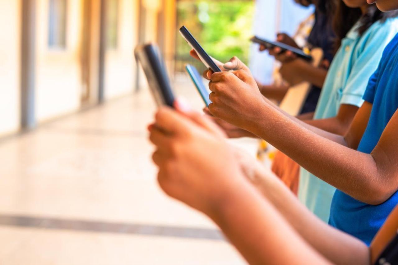 Photo shows a group of children using smartphones in a school corridor, highlighting the role of social media, gaming, technology, and education in their daily lives. (Adobe Stock Photo)