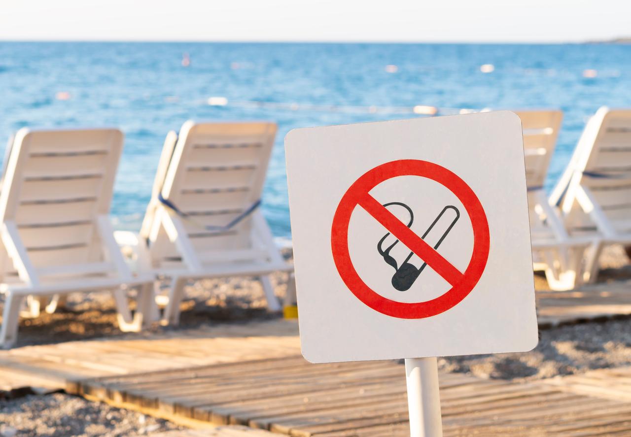 A no-smoking sign is placed on a beach near sun loungers facing the sea, indicating a designated smoke-free area along the coastline. (Adobe Stock Photo)