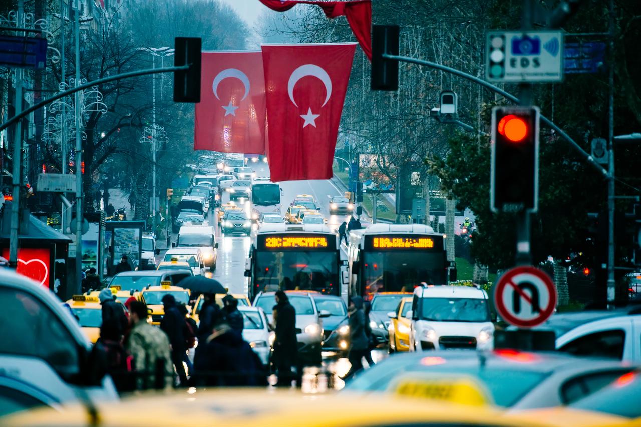 Traffic a rain-slicked avenue in Istanbul, where brake lights and headlights reflect off wet asphalt under Turkish flags. (Photo via Adobe Stock)