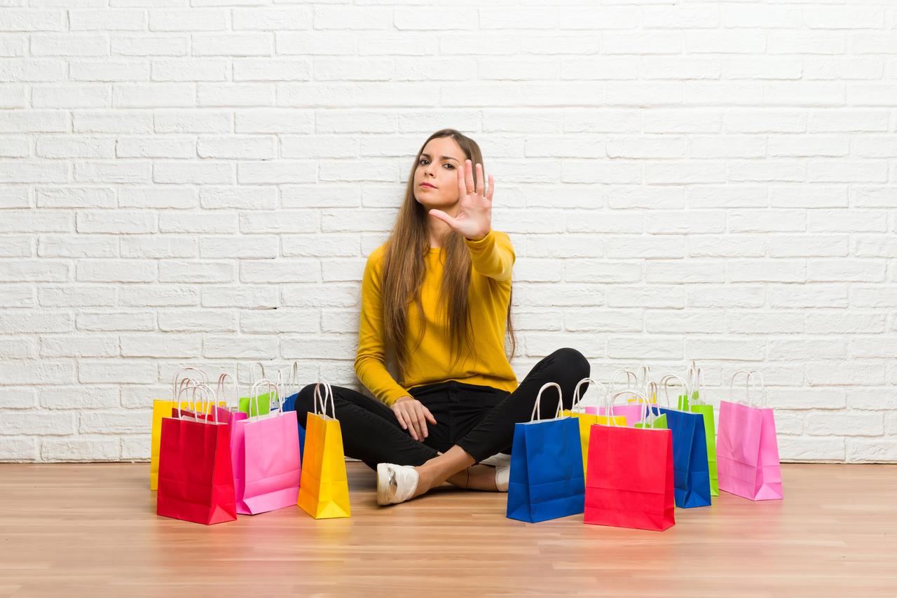 A woman sits among shopping bags while raising her hand in a stop gesture, illustrating resistance to excessive shopping or consumerism. (Adobe Stock Photo)