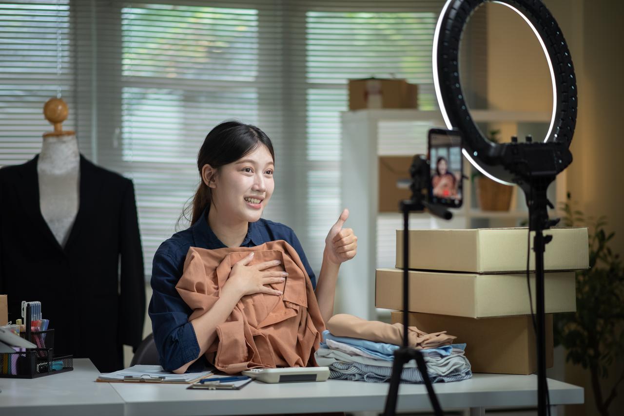 A woman presents clothing during a livestream sales session using a smartphone and a ring light. (Adobe Stock Photo)