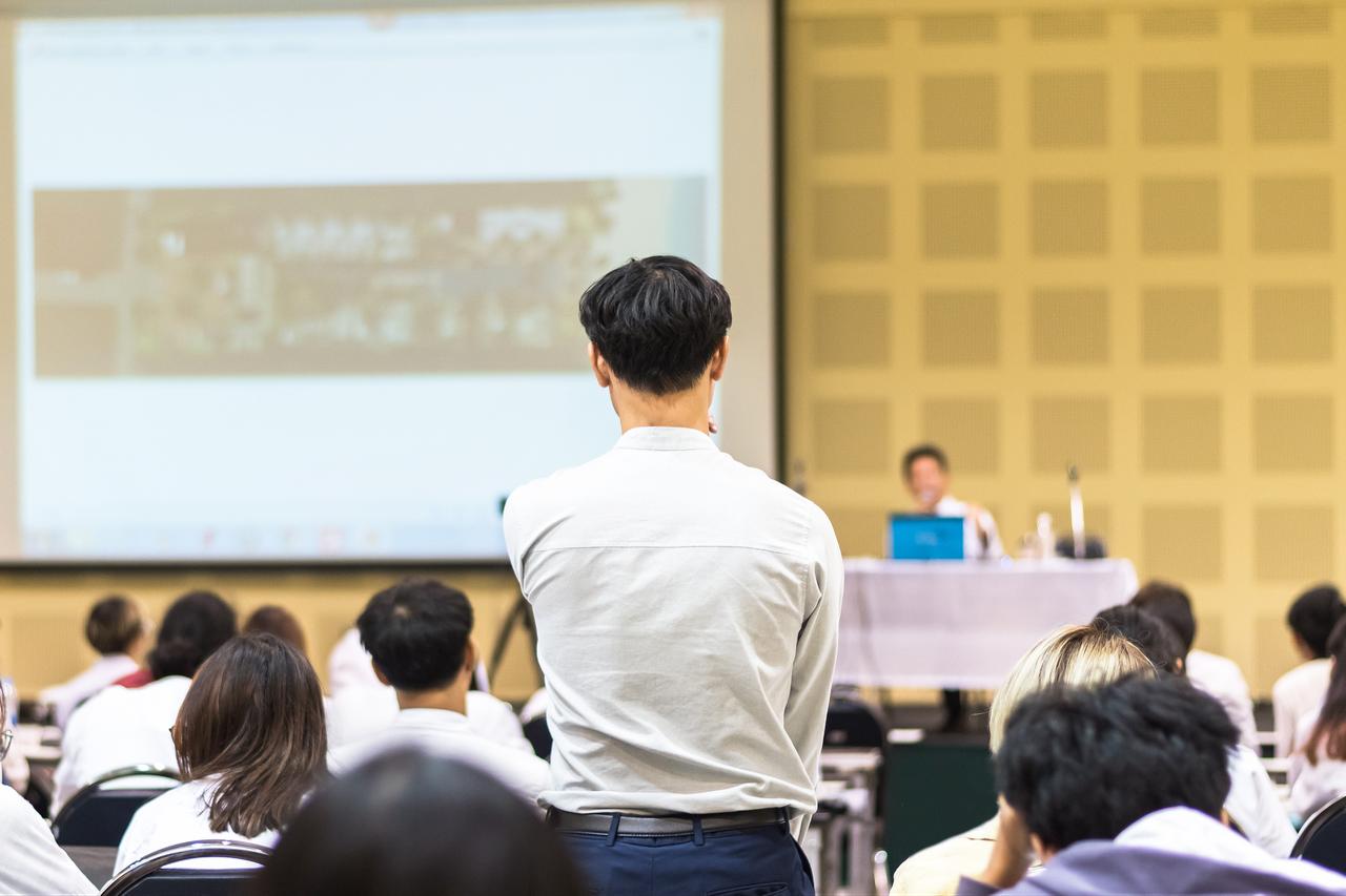 A participant stands to ask a question during a conference or seminar attended by a seated audience. (Adobe Stock Photo)