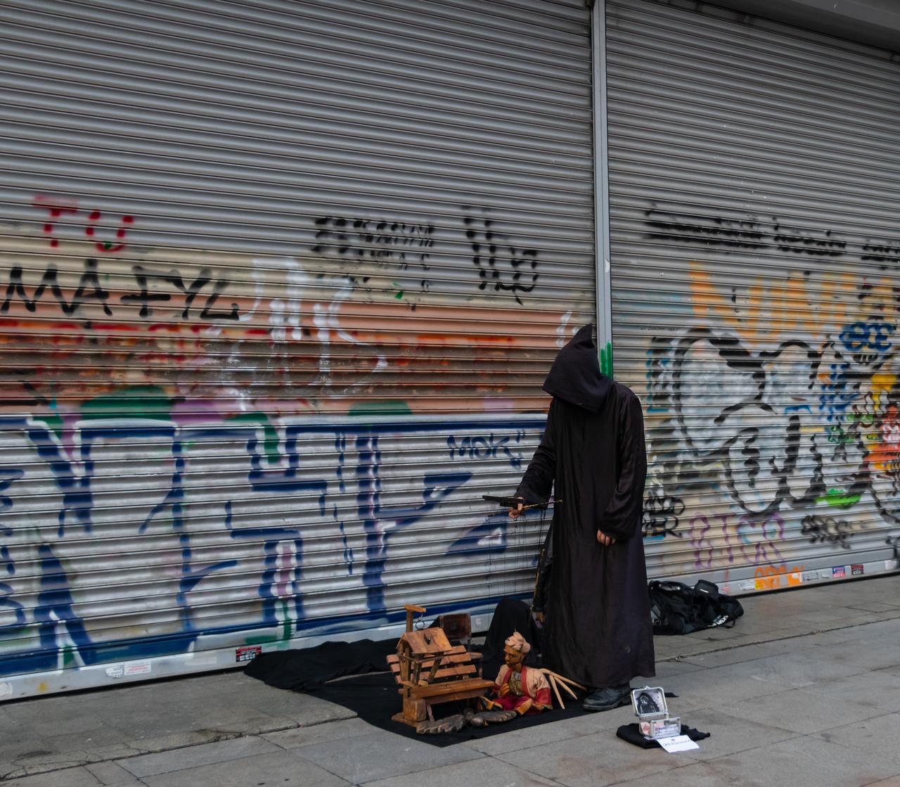 A street performer dressed in a dark costume stands in front of a graffiti-covered metal shutter. (Adobe Stock Photo)