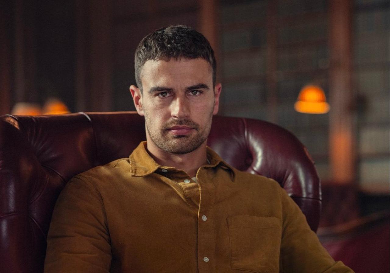 Theo James wearing a brown shirt sits in a leather armchair. (Photo via social media)