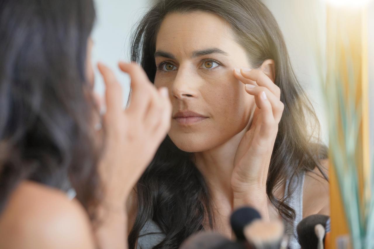 A woman examines the skin around her eyes while looking in a mirror. (Adobe Stock Photo)