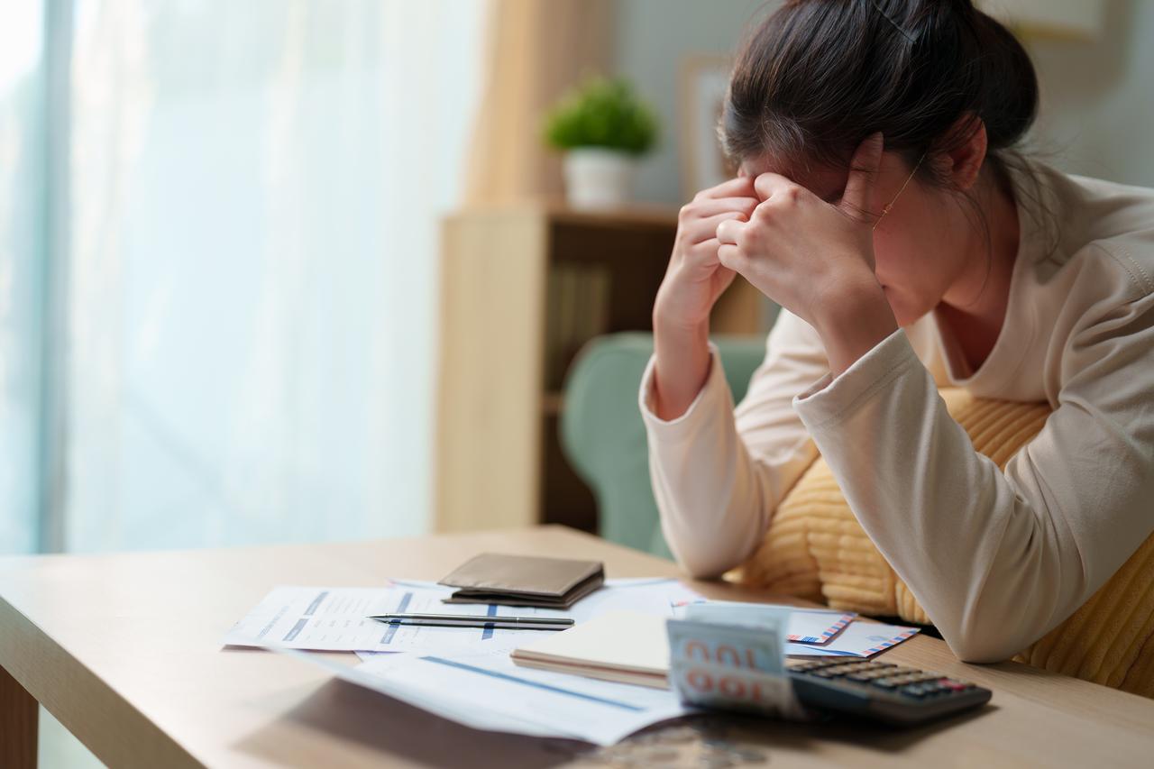 A woman holds her head while sitting at a table with bills, financial documents and a calculator. (Adobe Stock Photo)